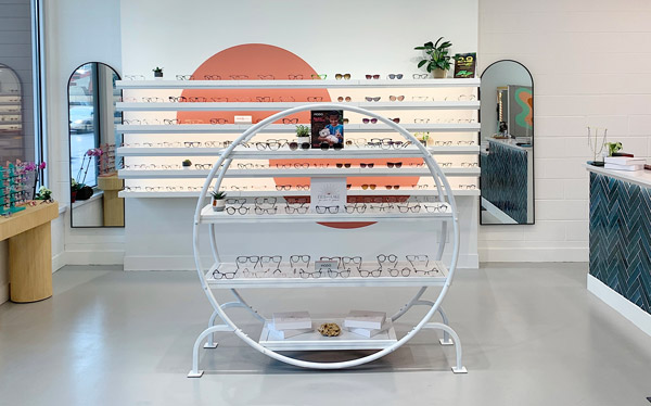 A creative frame board display in the office of Dr. Vaugn Schneider, who was recently featured on Women In Optometry. The display has a backlit wall with an orange paint feature and clean, white shelves of frames. In the foreground, a round, metal shelving unit also displays sunglasses and other eyewear.