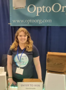 Elizabeth Hunt stands at a trade show booth under the optoorg banner. Company's new product reminds wearers to replace contact lens cases. 