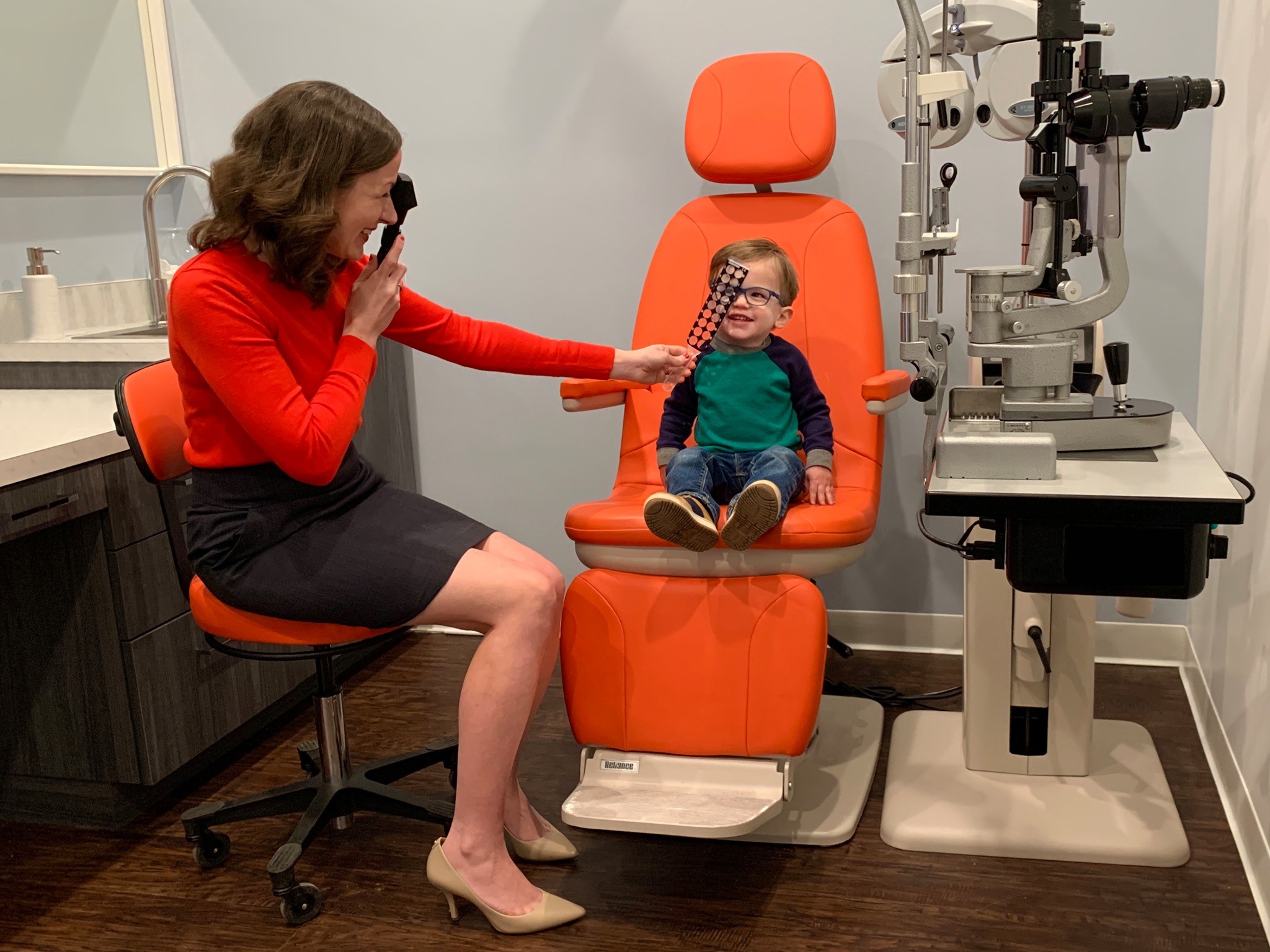 Dr. Lyons is seated examining a young child who sits in the big orange chair